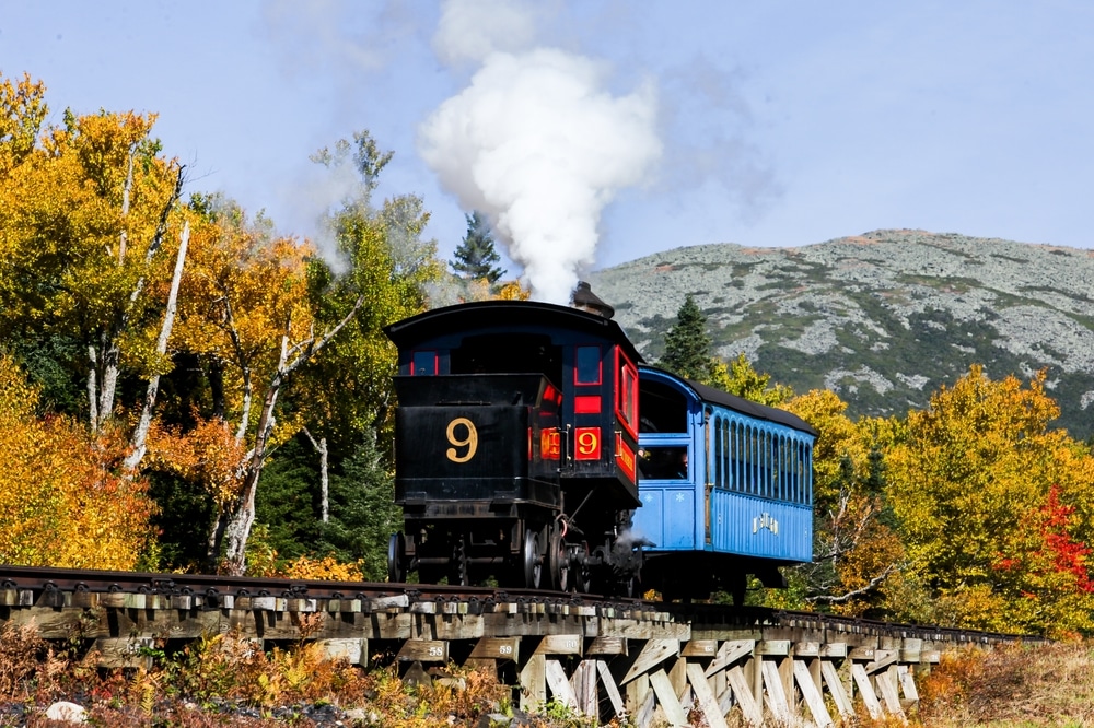 Ride the Mt. Washington Cog Railway this fall. Number 9 tug pulling a blue passenger rail car with fall foliage in the background.